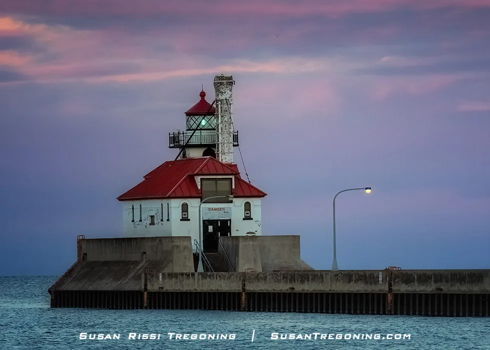  The Duluth South Breakwater Outer Light stands at the end of a concrete pier during a sunset with pink and purple tones in the sky. The lighthouse’s red roof and illuminated lantern room are visible, along with a single lit streetlamp on the pier. Calm water surrounds the pier beneath the colorful evening sky.