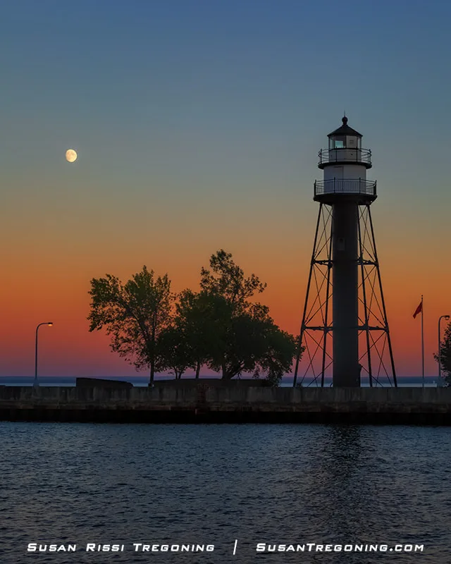 The Duluth Harbor South Breakwater Inner Light stands on a pier at dusk, silhouetted against a sky with deep blue, orange, and pink tones. The moon is visible to the left above the lighthouse, and a few trees line the pier. Calm water reflects the colors of the fading sunset.