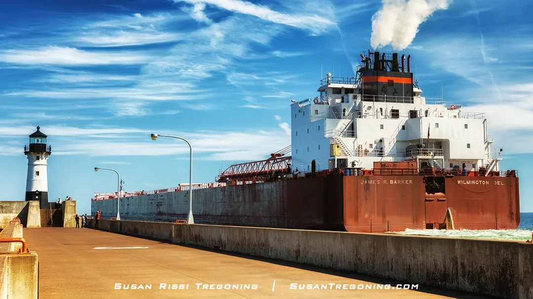 The James R. Barker passes the North Pier Light as she exits the Duluth Ship Canal, with people standing along the pier and a bright blue sky overhead.