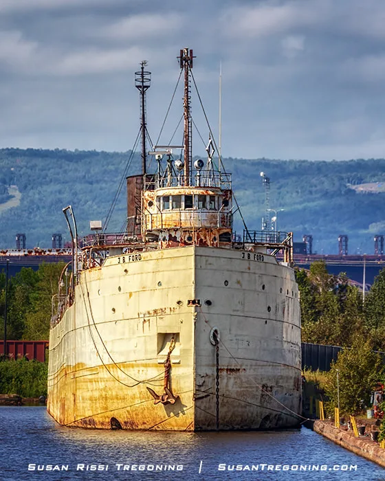 The weathered cargo ship J.B. Ford sits docked in a narrow waterway, its rusted bow facing the viewer with anchor chains and ropes hanging along the hull. The ship’s superstructure rises behind the bow with masts and antennas, and industrial buildings and a hilly landscape appear in the background under a partly cloudy sky.