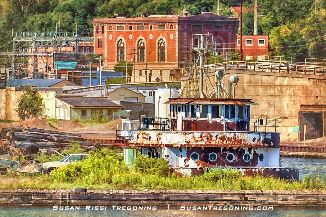 A rusted tugboat pilothouse sits along an overgrown waterfront, with industrial brick buildings, electrical equipment, and power lines behind it. The structure shows peeling paint and weathering, and the surrounding area appears aged and unused under partly cloudy light.