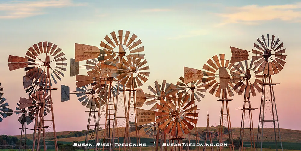 Several vintage metal windmills stand in a grassy field at sunset, each with weathered blades and tail vanes. Rolling hills and a colorful sky fill the background.