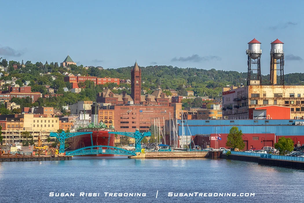  A view of the Minnesota Slip Drawbridge in Duluth, Minnesota, seen from the water. The bright turquoise double drawbridge spans the slip near the waterfront. Industrial buildings and two cylindrical water towers stand behind it, with historic brick structures rising up the hillside in the background under a partly cloudy sky.