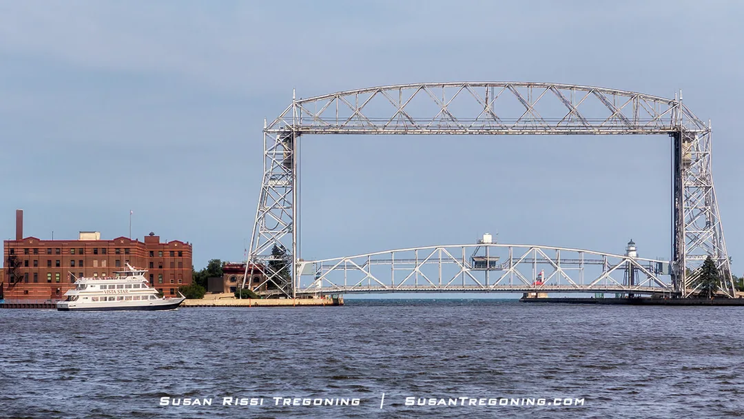 The Vista Star sightseeing boat sits in Duluth Bay near the Aerial Lift Bridge, with the steel bridge structure spanning the waterway and a brick building with a tall chimney on the left. The water reflects the boat and bridge, and the sky is clear and light blue.