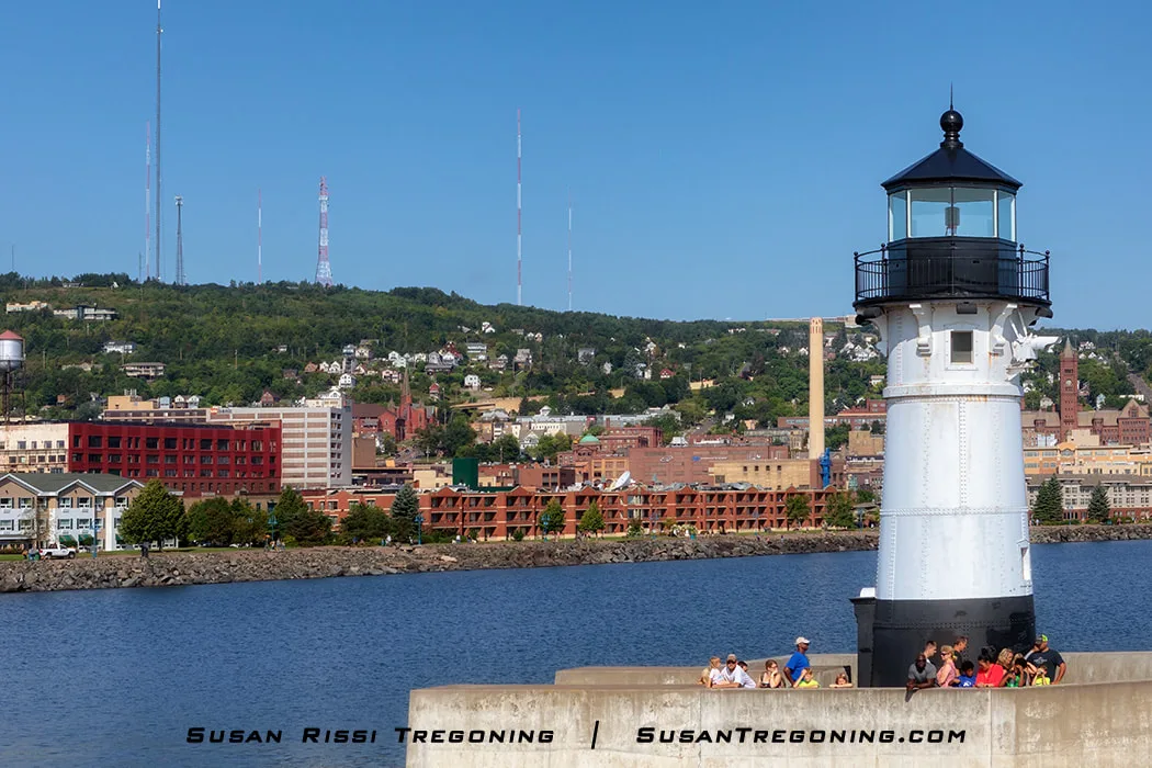  The Duluth North Pier Light stands on a concrete pier in bright daylight, with several people gathered near its base. Calm water fills the foreground, and the city of Duluth rises on the hillside behind the lighthouse, with buildings, houses, and tall communication towers visible against a clear blue sky.