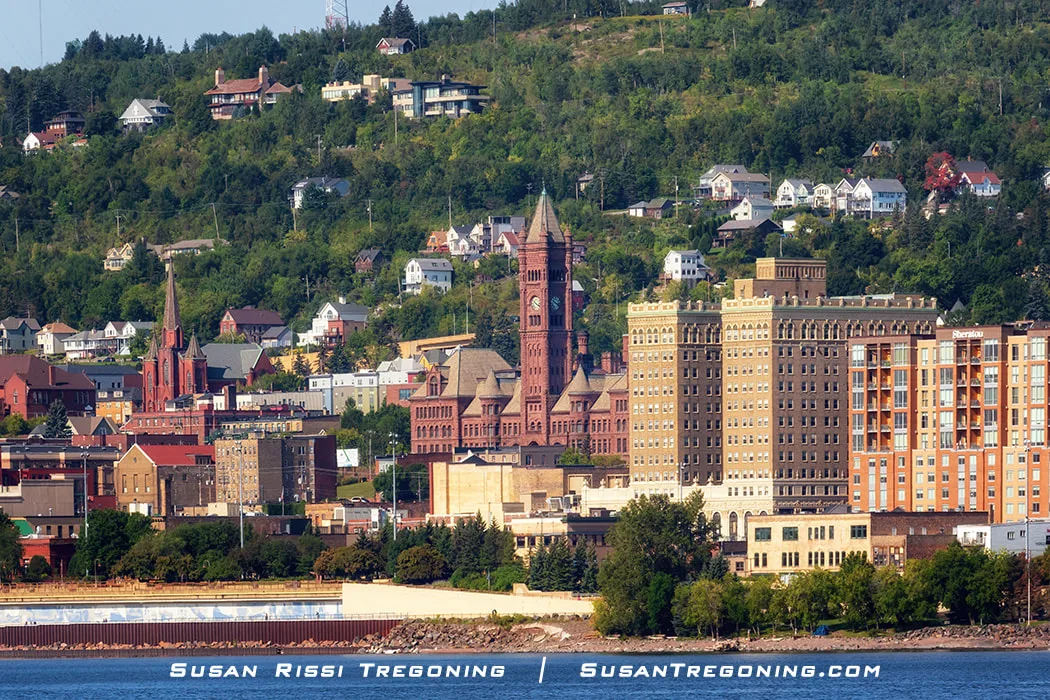  
A view of downtown Duluth, Minnesota, taken from across Lake Superior. The tall red‑brick clock‑tower building of Historic Central High School stands prominently on the hillside. Nearby, the Sacred Heart Cathedral rises with its pointed steeple. Modern buildings, including one with a Sheraton logo, appear in the foreground, while houses and trees cover the hillside behind the historic structures.