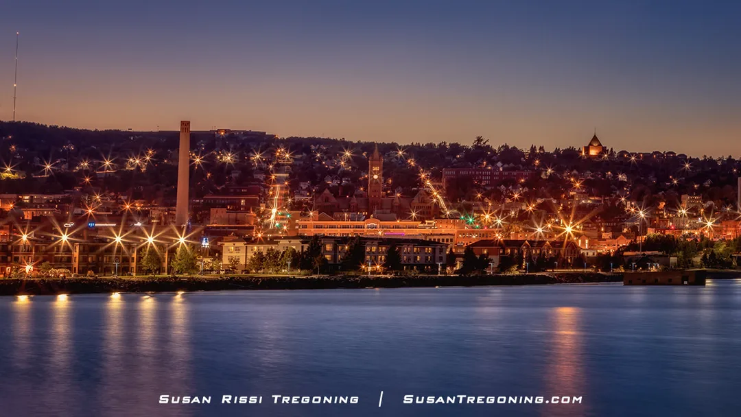 A nighttime view of Duluth, Minnesota, seen from across Lake Superior. City lights create starburst effects along the waterfront, reflecting across the calm water. Buildings rise up the hillside, including a tall smokestack and a clock‑tower structure near the center. The sky transitions from deep blue to lighter tones near the horizon.