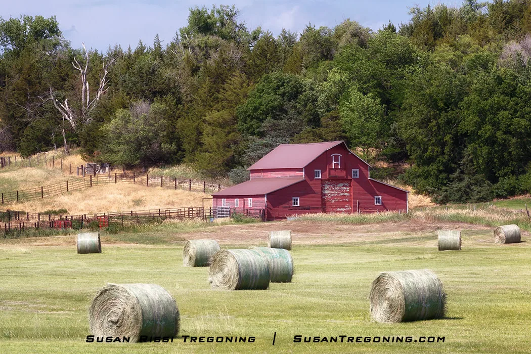 A red barn stands near a fenced area behind several large round hay bales scattered across a mowed field, with trees and rolling grassy hills in the background under a partly cloudy sky.
