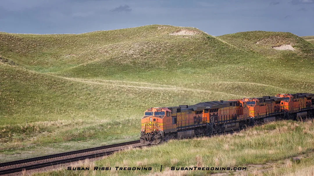A freight train with orange and black locomotives travels along railroad tracks that curve through rolling grassy hills under a partly cloudy blue sky.