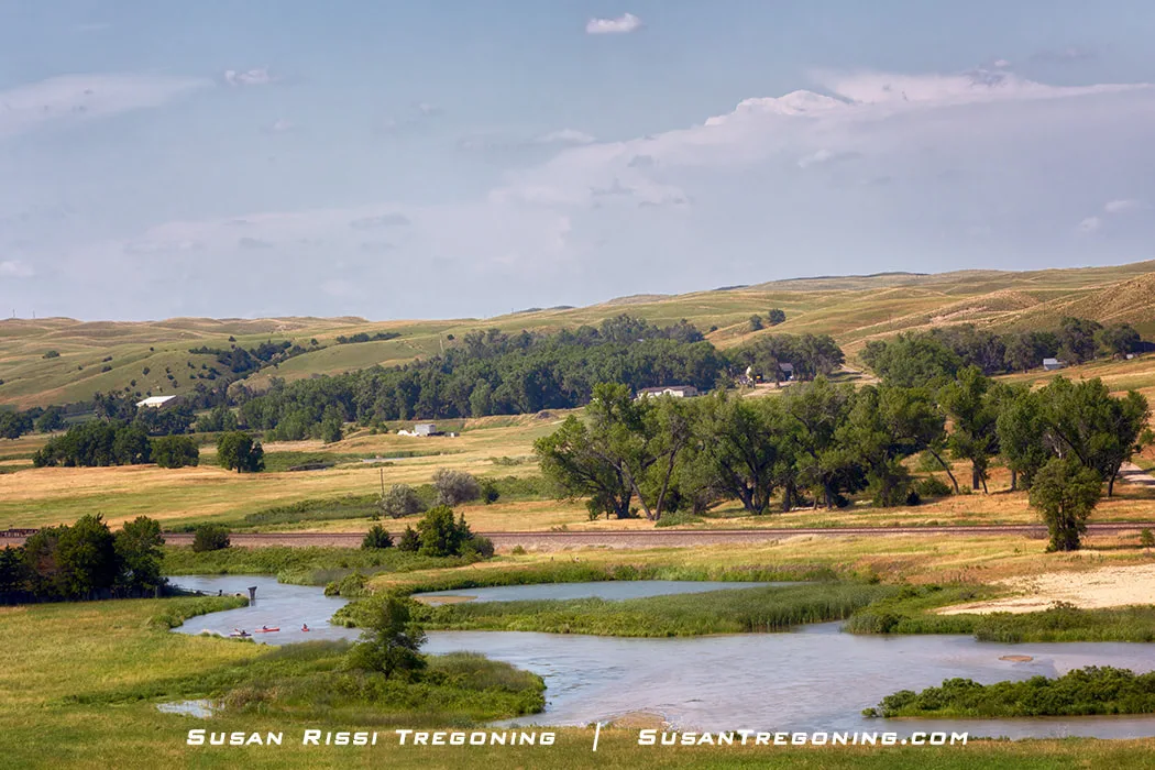 A winding river bordered by green vegetation flows through open fields and rolling hills under a mostly clear sky.