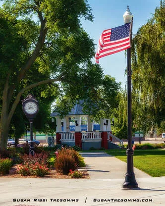 A park scene shows a lamppost with an American flag, a decorative street clock, and a gazebo surrounded by trees and landscaped flower beds. Paved walkways and green lawn areas fill the town square.