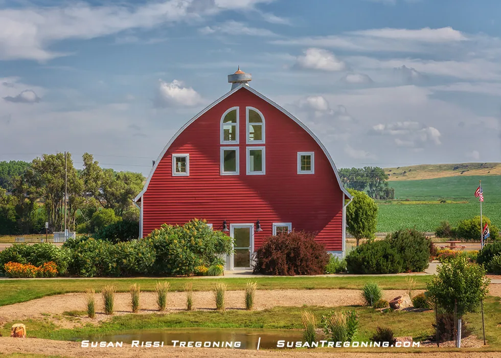 A bright red barn with white trim stands in a landscaped area with shrubs, small trees, and a gravel path. Green fields and rolling hills stretch into the distance beneath a partly cloudy blue sky.