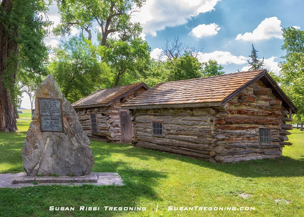 A small log cabin with a wood‑shingle roof stands in a grassy area surrounded by trees. A tall stone monument with a metal plaque is positioned in front of the cabin.
