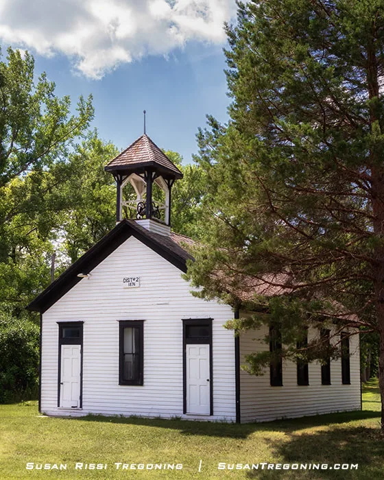 A white wooden one‑room schoolhouse with a bell tower stands on a grassy lawn. The front has two doors, three windows, and the sign “Dist. #2 1874.” Trees surround the building under a partly cloudy sky.
