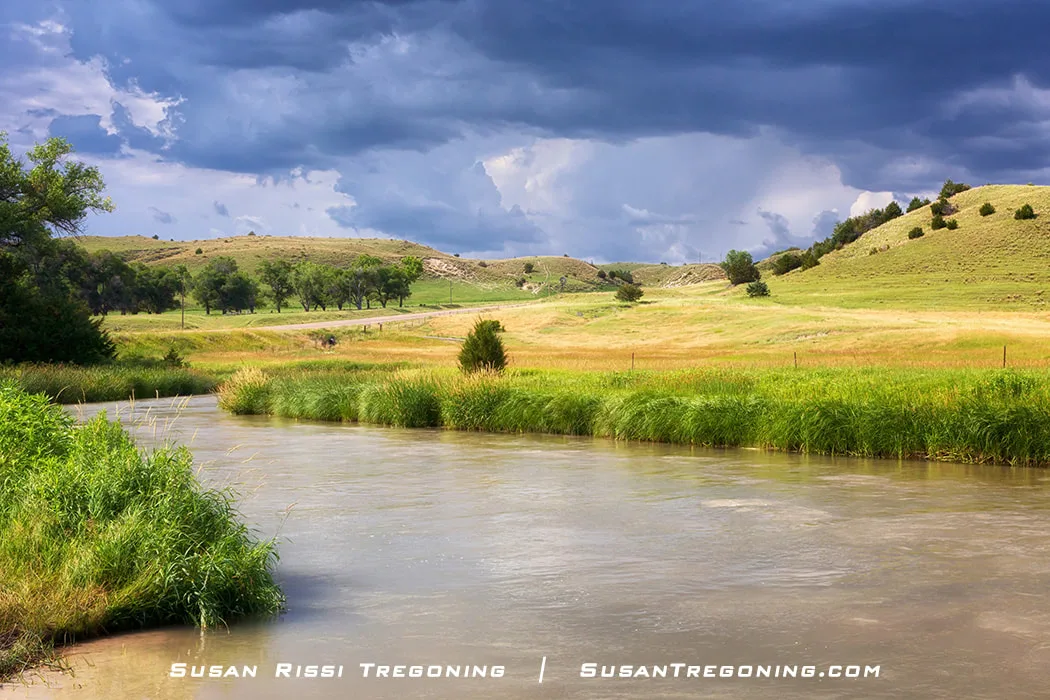 A river winds through rolling grassy hills under dark storm clouds, with sunlight illuminating the landscape in contrast to the approaching weather.