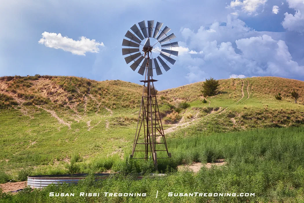 A tall metal windmill stands in a grassy area beside a round metal stock tank, with rolling hills and scattered shrubs in the background under a partly cloudy sky.