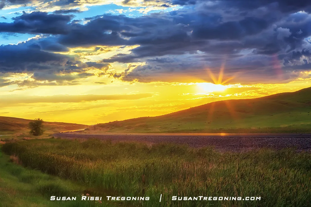 A sunset glows over rolling grassy hills, with the sun low on the horizon casting warm orange and gold light across the sky. In the foreground, railroad tracks curve through the landscape, reflecting the sunlight.