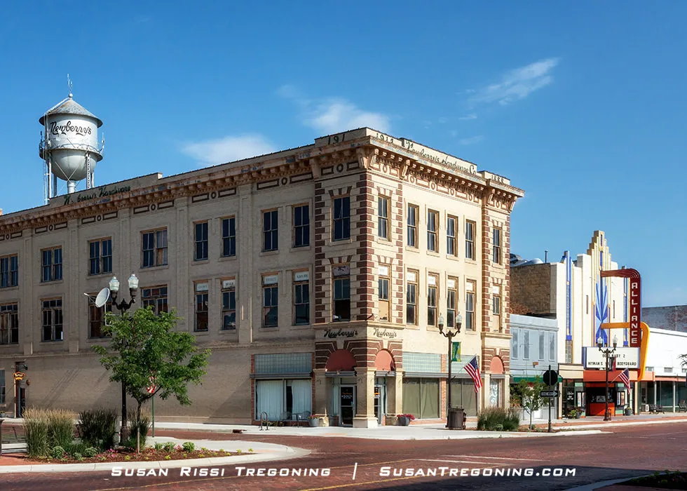A three‑story Neoclassical Revival brick building stands along a downtown street, with large first‑floor storefront windows and upper‑story windows marking the former office and workshop levels.