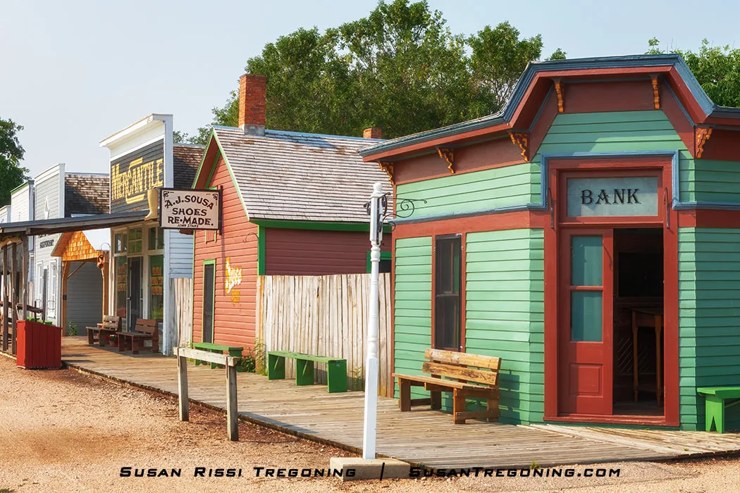  A row of colorful wooden storefronts lines a dirt street, including a green and red building labeled “BANK,” a red and green shop with a sign reading “A.J. Sousa Shoes Re‑Made,” and additional buildings extending down the boardwalk.