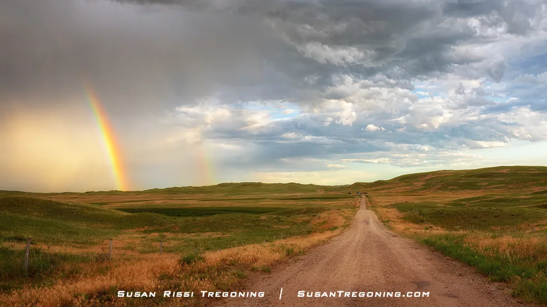 A dirt road stretches through rolling grassy hills beneath a partly cloudy sky. A bright rainbow arcs down to the left side of the scene, with a faint second rainbow beside it. A fence line and patches of golden grass fill the foreground.