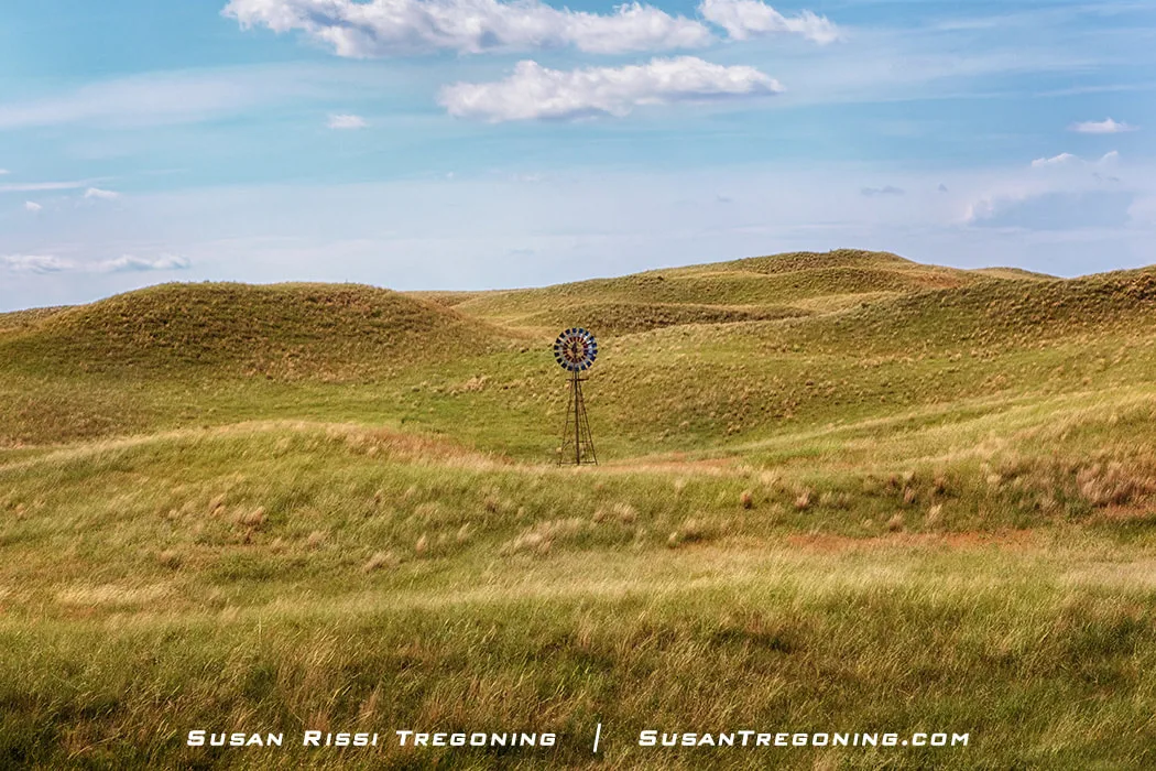 A windmill painted in red, white, and blue stands on a grassy hill surrounded by rolling golden‑green hills under a partly cloudy blue sky.