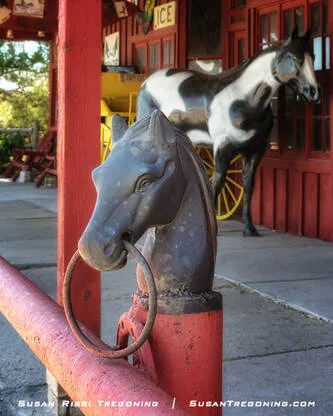 A cast‑iron horse‑head hitching post with a metal ring in its mouth is mounted on a red railing, with a life‑size horse figure and a rustic storefront in the background.