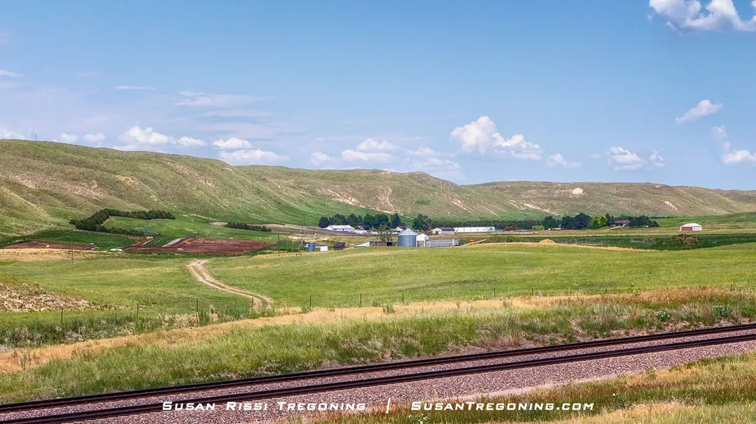 A wide rural landscape with rolling green hills, open fields, and a ranch in the middle distance. Several farm buildings and silos sit among the fields, with a dirt road curving toward them. Railroad tracks run across the foreground, and the sky above is bright blue with scattered white clouds.