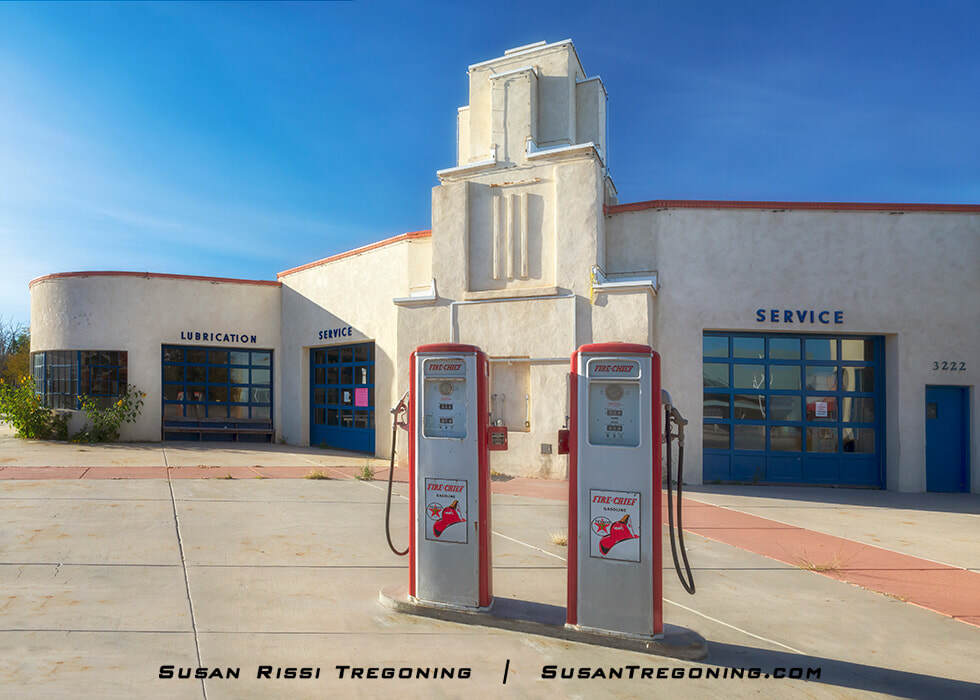 Two vintage Fire Chief gas pumps stand in front of a stucco service station with blue trim. The building features rounded Streamline Moderne styling, garage doors labeled “Lubrication” and “Service,” and the number 3222 on the right side. The scene is photographed in daylight.