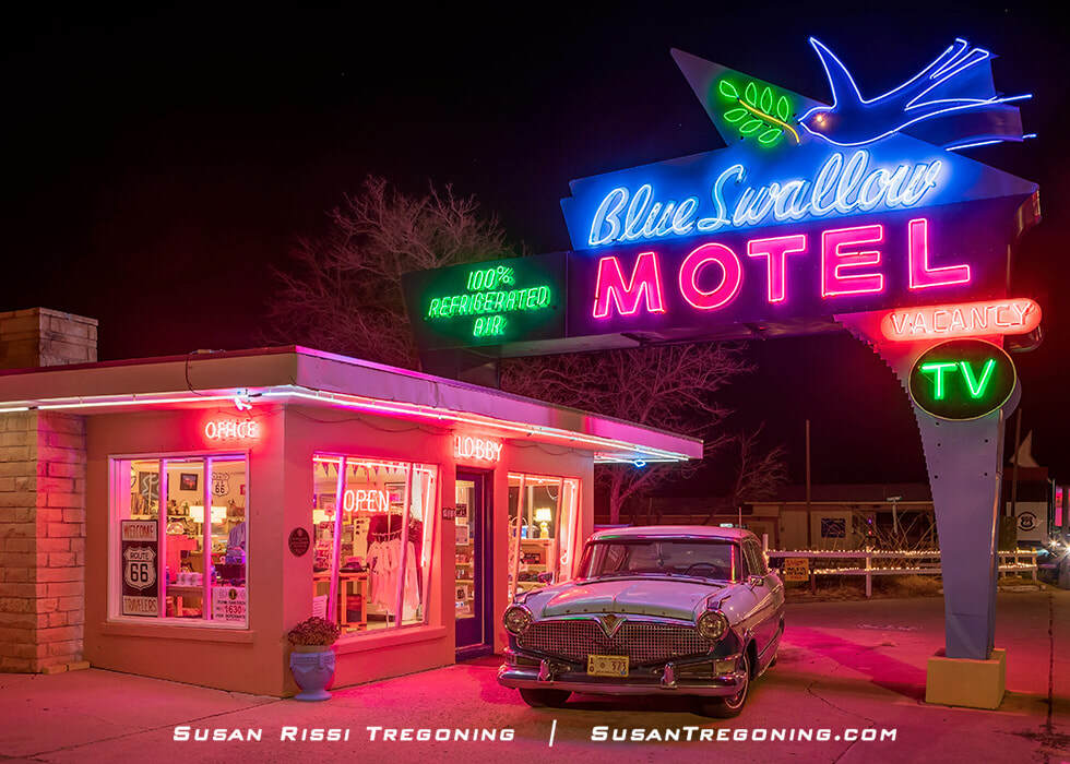 A nighttime view of the Blue Swallow Motel on Route 66, showing its brightly lit neon sign with a blue swallow graphic and the words “Blue Swallow Motel.” The office building is illuminated with additional neon accents, and a classic car is parked in front of the lobby.