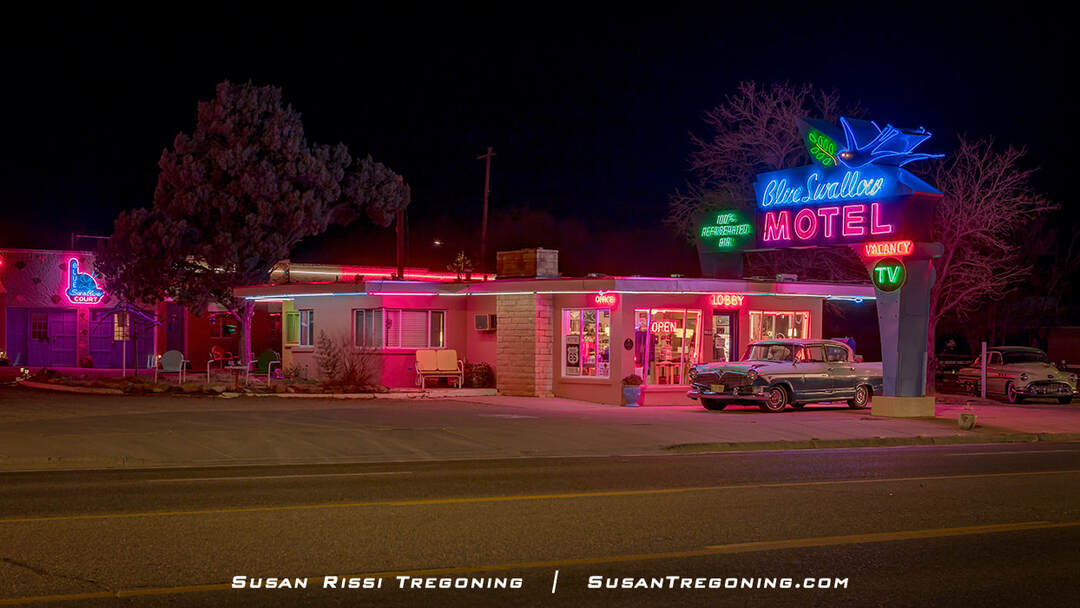 A nighttime view of the Blue Swallow Motel in Tucumcari, New Mexico, with its neon signs fully lit, including the main “Blue Swallow Motel” sign, smaller vacancy and amenity signs, and additional neon accents on the building. A classic car is parked in front of the lobby, and the stone‑front motel façade is illuminated by the glow.