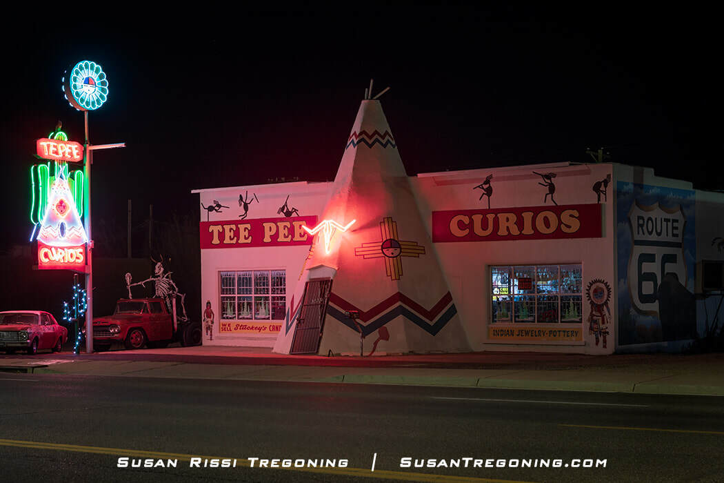 A nighttime view of Tee Pee Curios in Tucumcari, New Mexico, showing the neon‑lit teepee‑shaped storefront and the vertical “Tepee Curios” sign. Painted designs and a large Route 66 shield mural appear on the building, with two vintage cars parked in front.