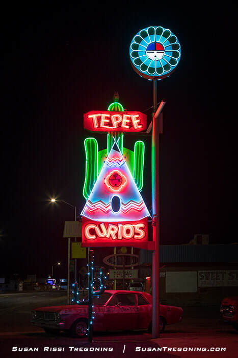 A nighttime view of the neon sign for Tepee Curios in Tucumcari, New Mexico, featuring a cactus, a teepee outline, and bold red lettering. The sign, added in the mid‑1960s, glows against the dark sky with a red vintage car parked below.