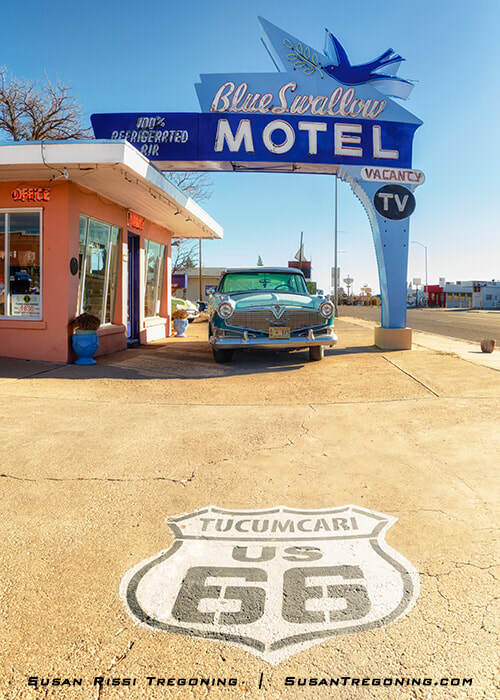 A daytime view of the Blue Swallow Motel in Tucumcari, New Mexico, showing its neon sign above the office and a classic car parked in front. The Route 66 shield with “Tucumcari US 66” is painted on the pavement in the foreground.