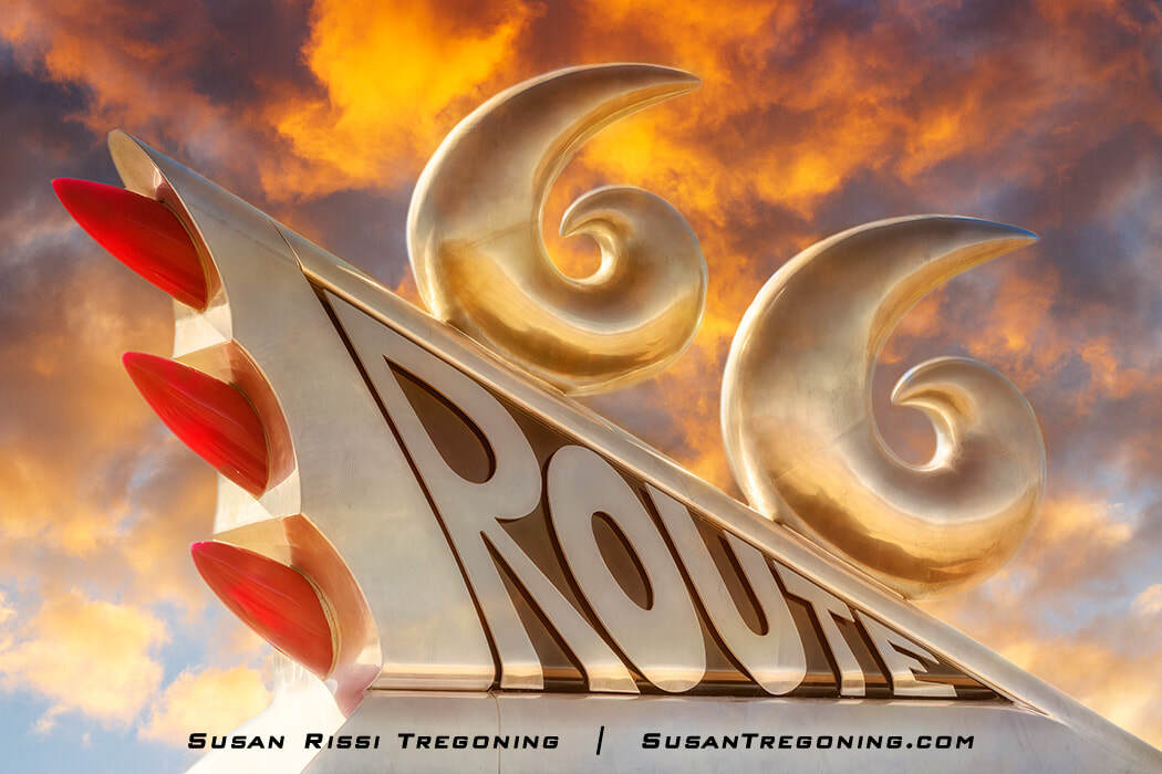 A chrome 1950s‑style tailfin sculpture with red bullet lights and two stylized sixes mounted at the top of the Route 66 monument, shown at sunset with warm light reflecting off the metal.