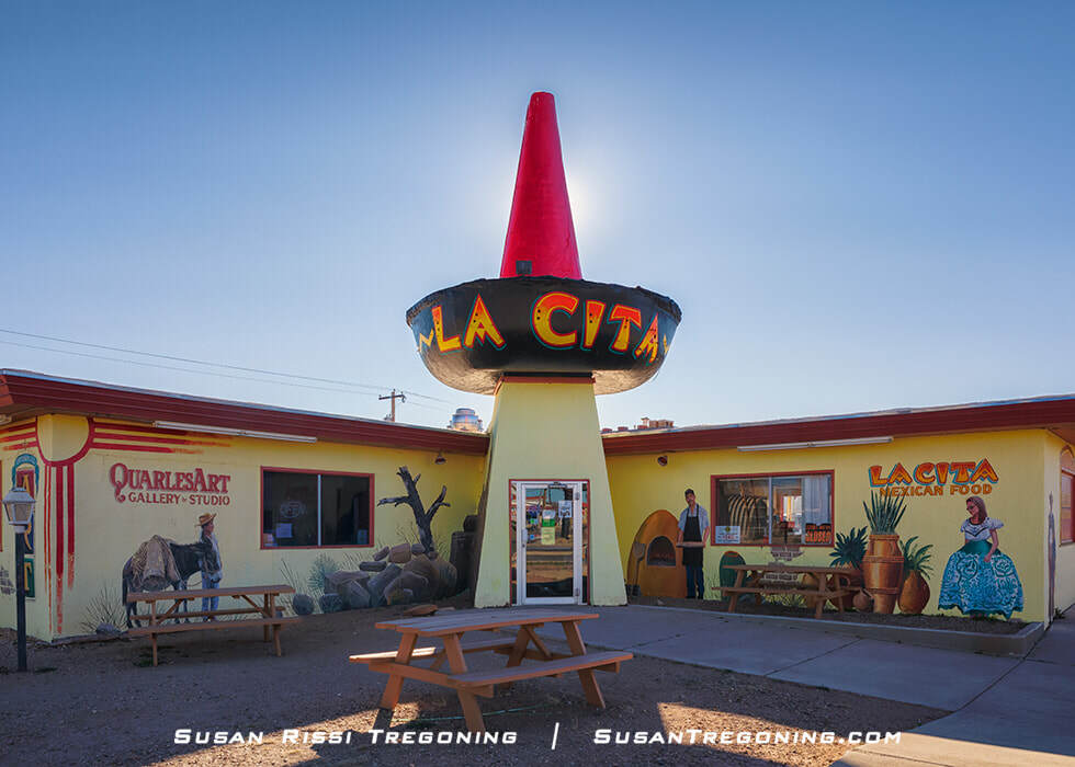 A brightly painted building in Tucumcari, New Mexico, with a large sombrero‑shaped structure on the roof labeled “La Cita.” Murals decorate the exterior walls, and picnic tables sit in front of the building with the sun positioned behind the sombrero.