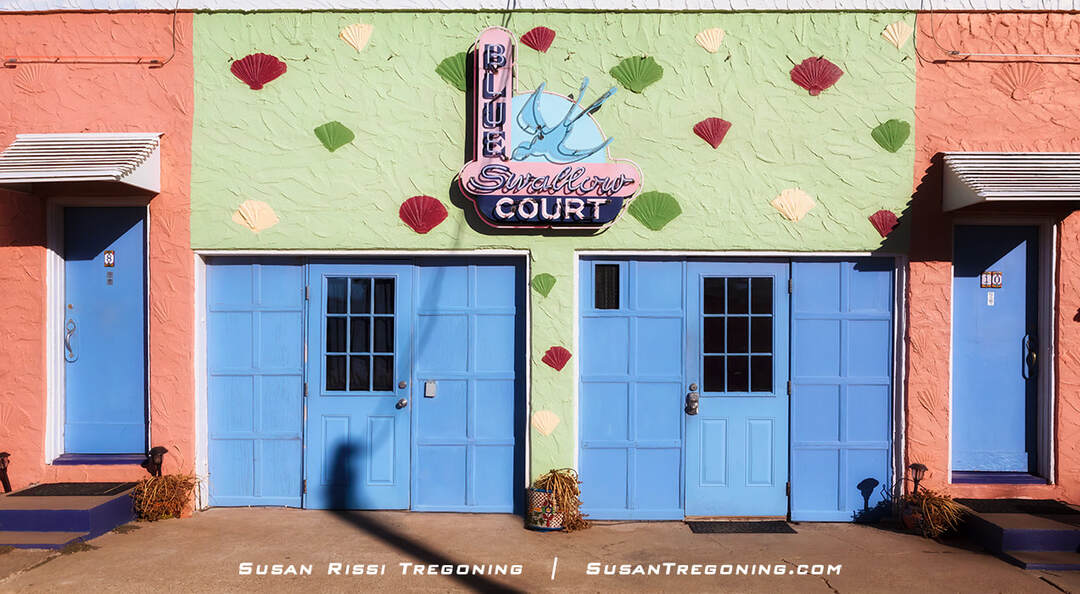 The original Blue Swallow Court neon sign from 1942, now mounted between two blue garage doors at the back of the Blue Swallow Motel. The pastel‑colored building façade features decorative shell‑shaped ornaments, and smaller blue doors with white awnings appear on either side of the sign.