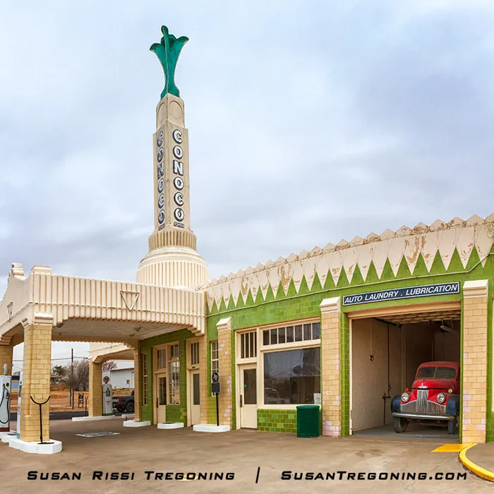 A restored Art Deco Conoco gas station in Shamrock, Texas, with a tall tower displaying the word “CONOCO,” cream and green geometric detailing, and an attached service bay labeled “Auto Laundry • Lubrication,” where a red vintage truck is parked.