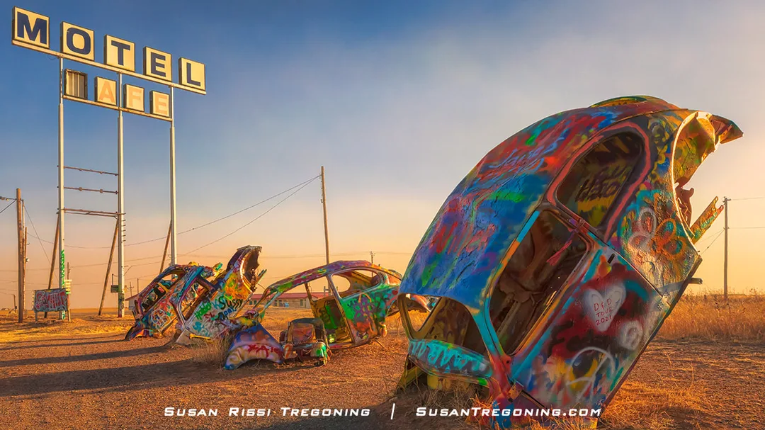 A row of partially buried Volkswagen Beetles at the Bug Ranch in Conway, Texas, each car covered in colorful graffiti and positioned nose‑down in the ground, with a weathered roadside motel and café sign and a warm sunset sky in the background.
