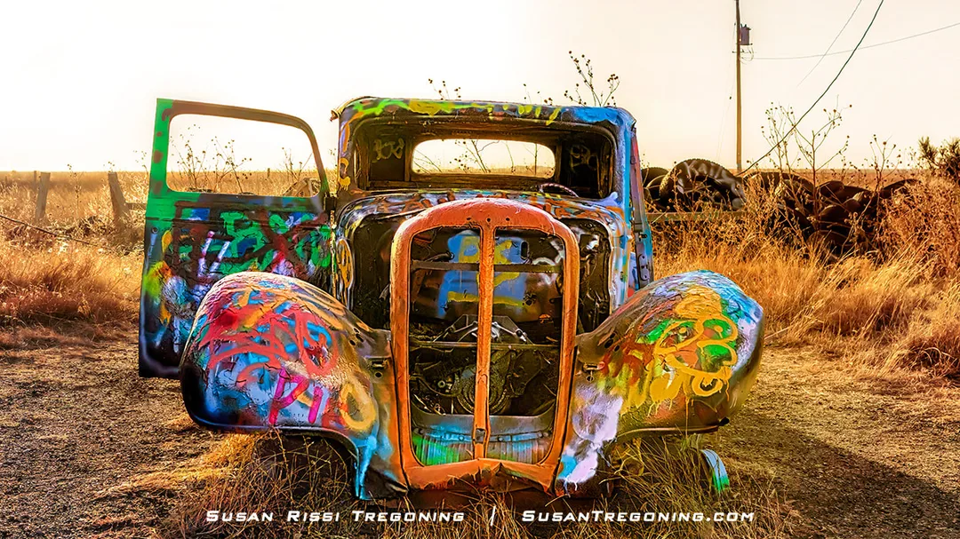 An abandoned vintage truck covered in colorful graffiti sits in a dry grassy field at sunset, with its doors open, rusted body exposed, and a wooden fence and utility pole in the background.