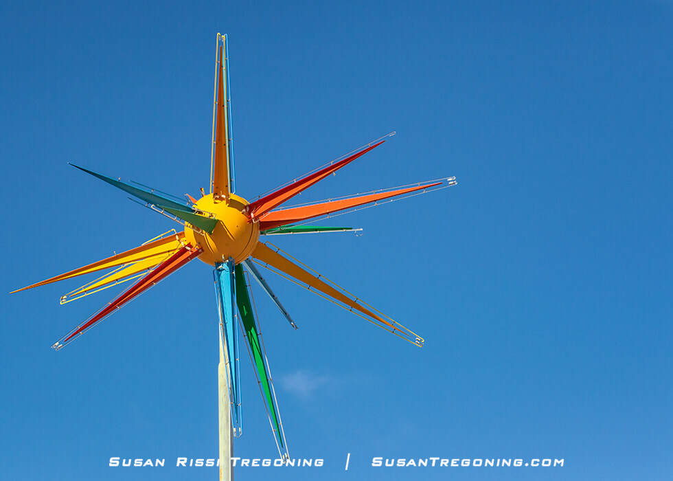 A colorful Roto‑Sphere sign in Moriarty, New Mexico, featuring a central yellow sphere with multiple glossy, triangular rays in orange, yellow, green, and blue extending outward like a sputnik‑style starburst. The sculpture is mounted on a tall pole against a clear blue sky.