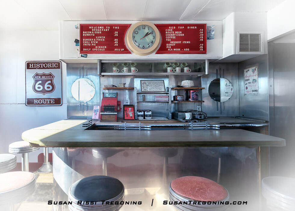 The interior of the Red Top Diner near Route 66 in Edgewood, New Mexico, showing a vintage lunch counter with round stools, chrome details, and a retro clock above the counter. A “Historic Route 66 – New Mexico” sign hangs on the wall, and shelves behind the counter hold mugs, condiments, and diner items. A menu board lists food and drink prices, all set within a classic mid‑century diner setting.