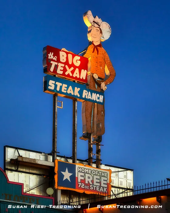 The brightly lit Big Texan Steak Ranch sign at dusk, featuring a cowboy figure above illuminated text advertising the restaurant and its “Home of the Free 72oz Steak” challenge against a deep blue evening sky.