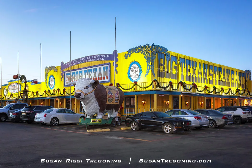 The exterior of the Big Texan Steak Ranch, a bright yellow and blue building decorated with lights and garlands, with large signs above the entrance and a white‑and‑brown bull statue displayed on a trailer in the parking lot, with several cars parked in front under a clear sky.