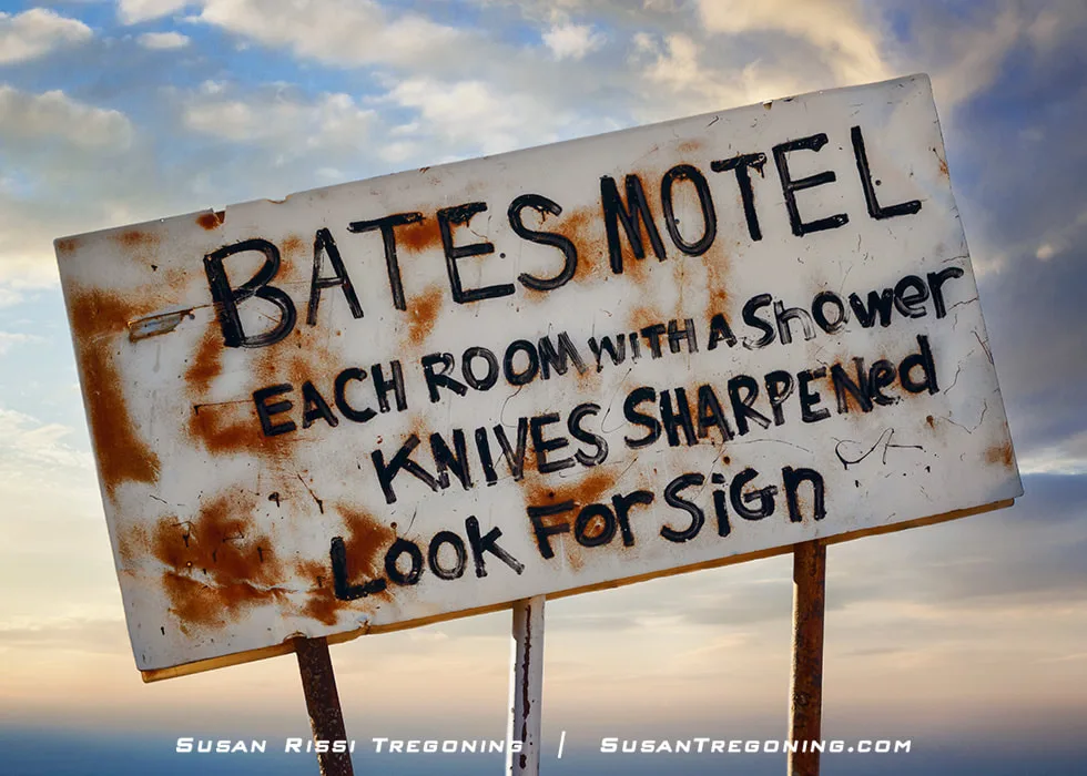 A weathered sign reading “Bates Motel, Each Room With a Shower, Knives Sharpened, Look For Sign” stands on two wooden posts against a cloudy sky, with rust stains and worn paint visible on the surface.