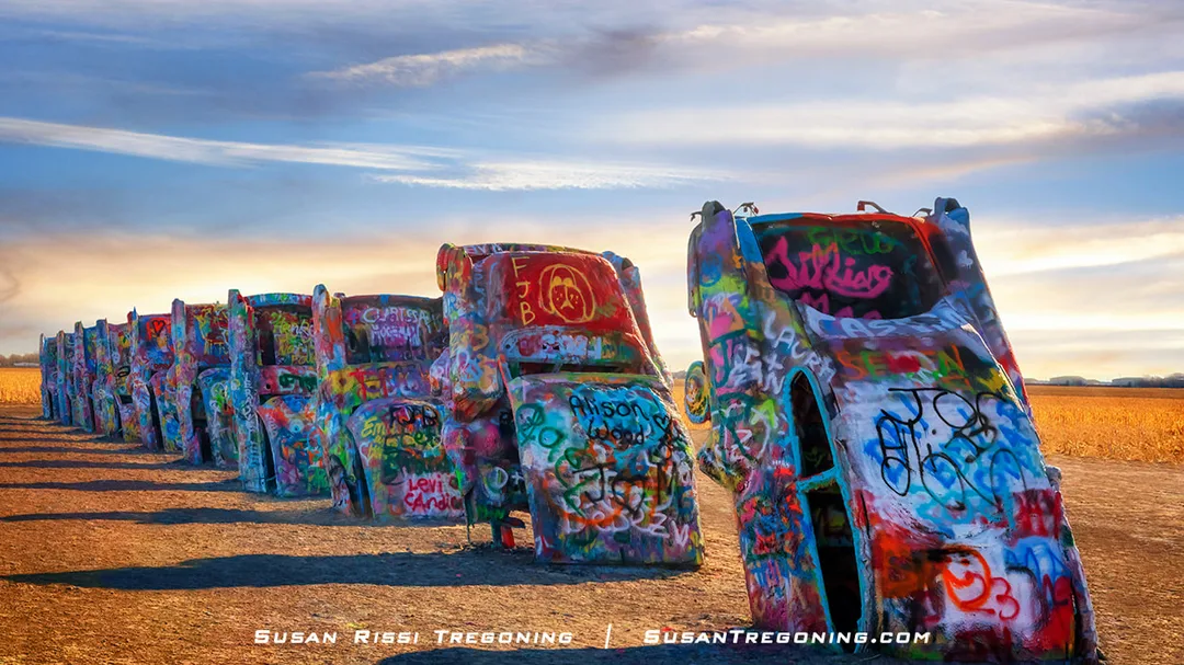 A row of half‑buried cars standing nose‑down in an open field, each one covered in colorful graffiti and spray paint beneath a partly cloudy sky.