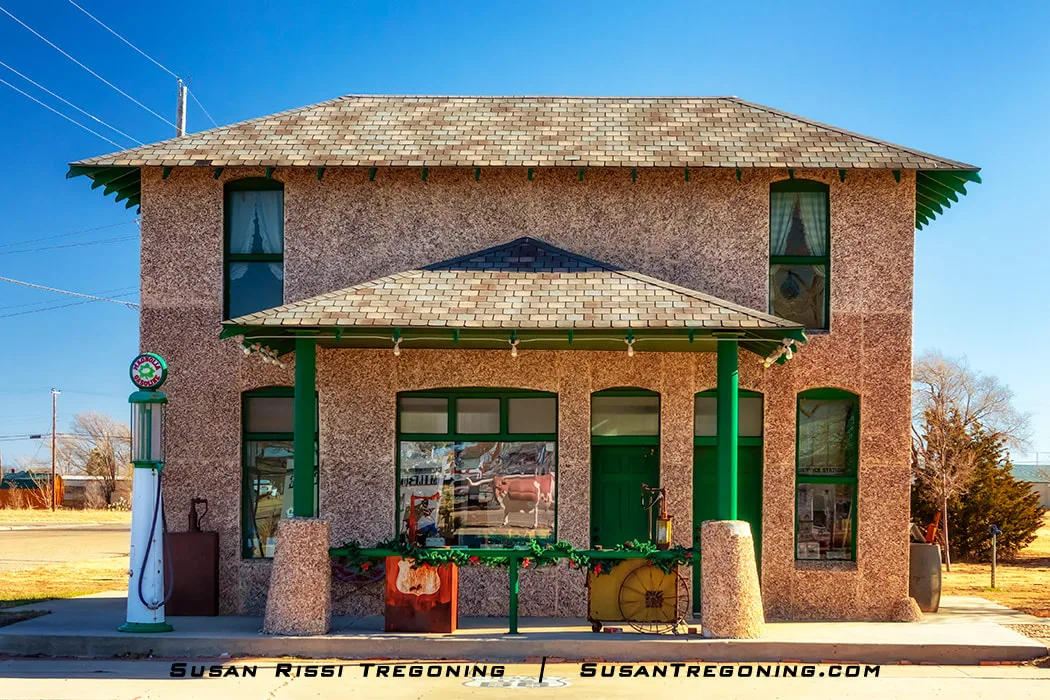 The restored Hiway Service Station on Route 66 in Vega, Texas, with a textured two‑story building featuring green trim, a vintage gas pump out front, and decorative items arranged near the entrance under a clear sky.