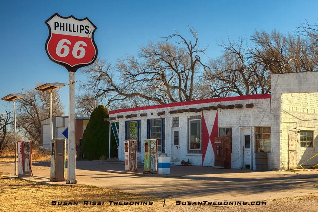 The Phillips 66 Midway Service Station in Adrian, Texas, with a tall Phillips 66 shield sign in front, a white brick building with red trim and an “X” pattern on the wall, and several rusted vintage gas pumps standing beneath a clear blue sky.