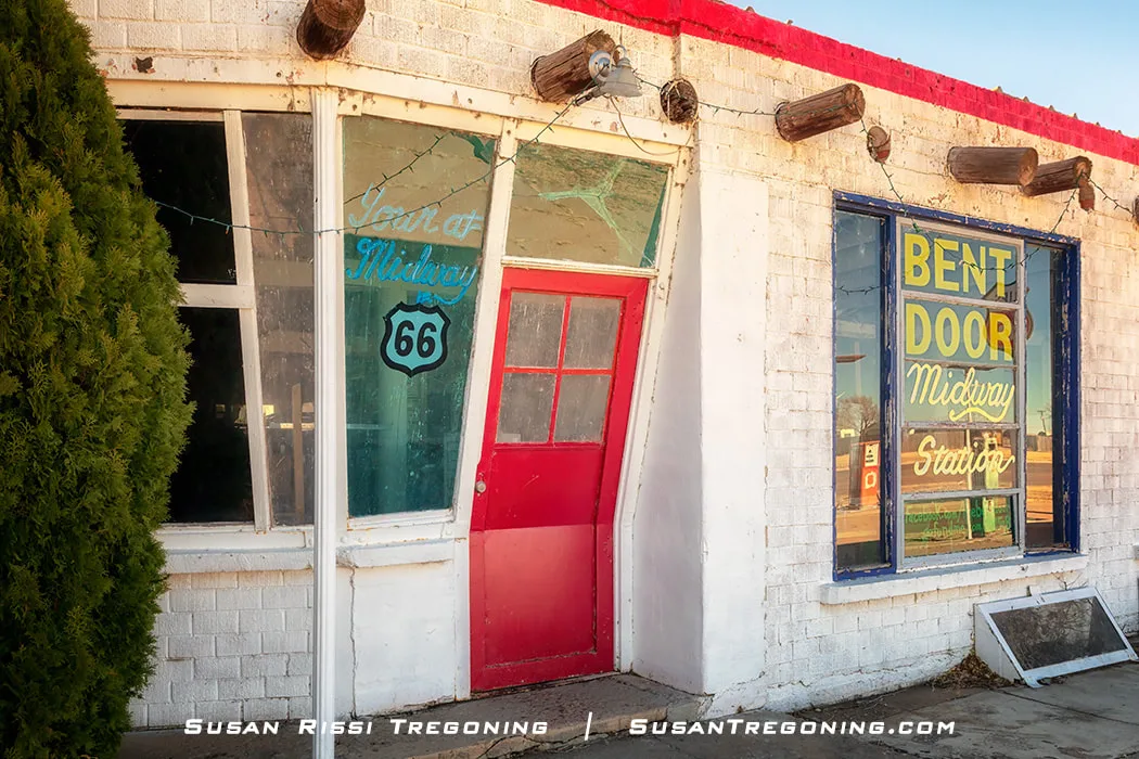 The Bent Door Café in Adrian, Texas, with a slanted red door on a white brick building, large windows displaying colorful Midway Station signage, red trim along the roofline, and string lights hanging across the front.