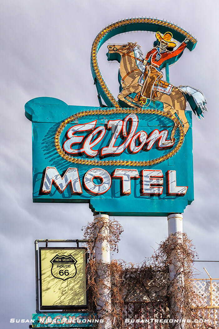 A vintage roadside motel sign with a turquoise background reading “El Don Motel.” Above the text is an illustrated cowboy on a rearing horse outlined with small bulbs. A smaller yellow sign below reads “New Mexico Route 66.” Vegetation partially surrounds the base of the sign.