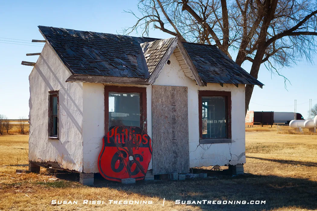   
Knox’s Phillips 66, a small white stucco building with a weathered shingle roof, two front windows, and a boarded‑up doorway, with a large vintage red and black Phillips 66 sign leaning against the wall in an open field with dry grass and scattered equipment in the background.